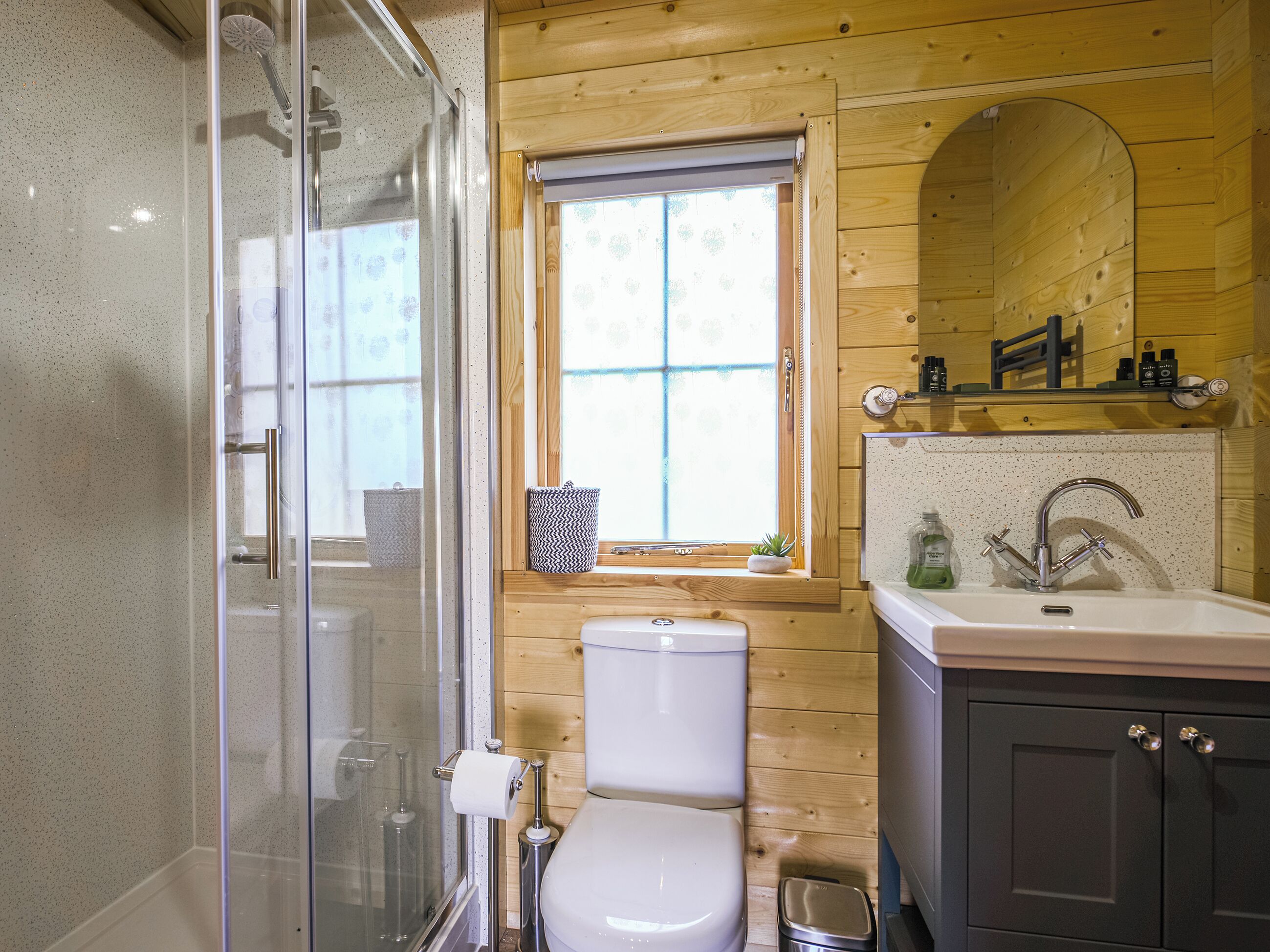 A bathroom with a glass shower enclosure, a toilet, a sink with a mirror, and a window with a view of greenery.