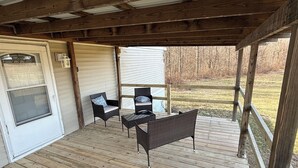 A covered porch with wooden flooring, two wicker chairs with cushions, a small table, and a view of a yard with trees. - Private Shawnee National Forest Retreat , Near Garden of the Gods (Elizabethtown)