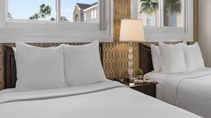 A hotel room with two beds, a nightstand with a lamp, and a view of buildings through the window.