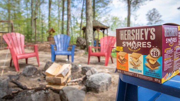 Une boîte de Hershey's S'mores sur une table, avec des chaises Adirondack et un feu de camp en arrière-plan.