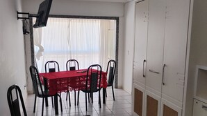 A dining area with a red table, black chairs, and a TV mounted on the wall. There is a large wardrobe with multiple doors and a ceiling light. - Vista da Orla de Itapoã - 1002 (Itapuã Vila Velha)