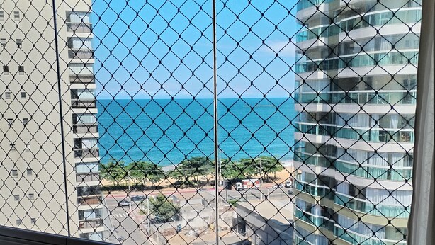 A view through a chain-link fence of a coastal area with high-rise buildings and a clear blue sky. - Vista da Orla de Itapoã - 1002 (Itapuã Vila Velha)