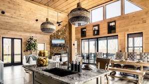 Una cucina moderna con un'ampia isola, un piano di lavoro in granito e una schiena a parete in pietra. Si vedono lampade a sospensione, un forno integrato e un'ampia finestra che lascia entrare la luce naturale.