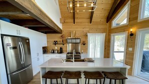 A modern kitchen with a stainless steel refrigerator, a central island with bar stools, wooden panel walls, and a large window. - Boutique chalet—woodland retreat, fireplace, chef kitchen, soaking tub—near PTDN (Val-Morin)