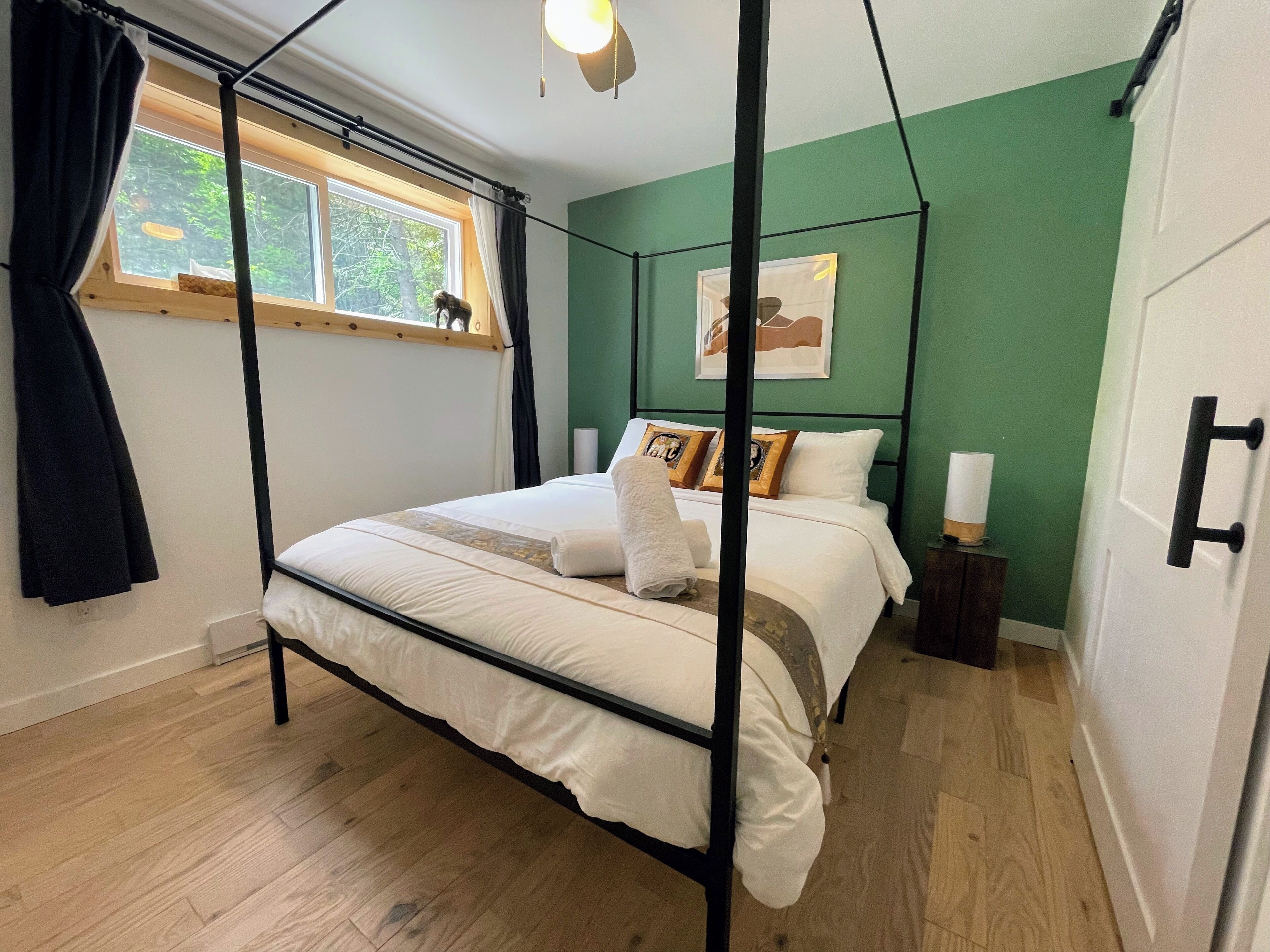 A four-poster bed with a white comforter and pillows, a bedside table with a lamp, and a window with curtains.