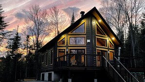 A modern log cabin with a large deck, multiple windows, and a staircase leading to the entrance. - Boutique chalet—woodland retreat, fireplace, chef kitchen, soaking tub—near PTDN (Val-Morin)
