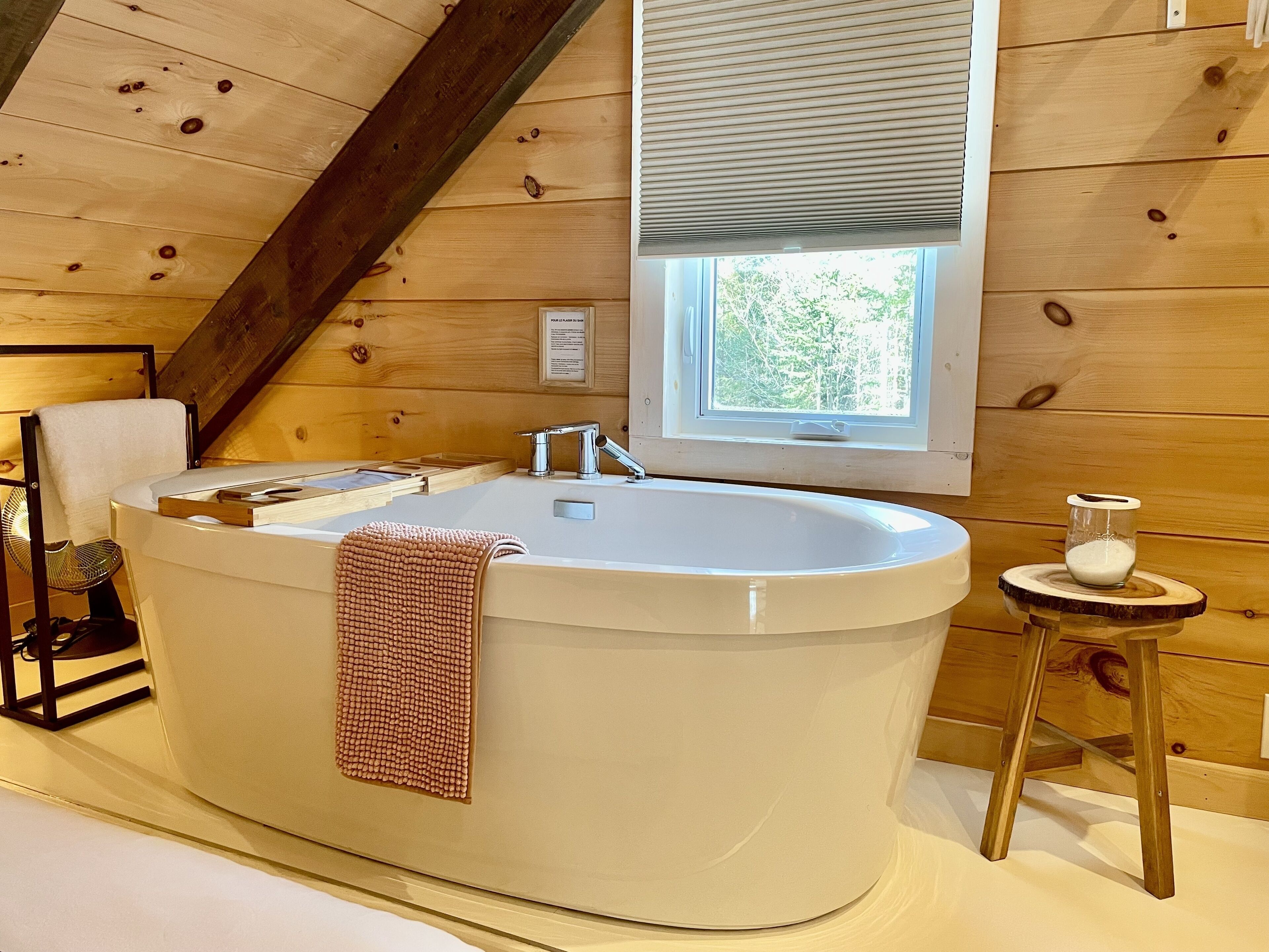 A modern bathroom with a freestanding bathtub, a window with blinds, and a small wooden stool with a glass container.
