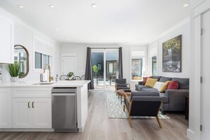 Una cocina moderna con armarios blancos, lavavajillas integrado y una zona de comedor con sofá gris y alfombra estampada.