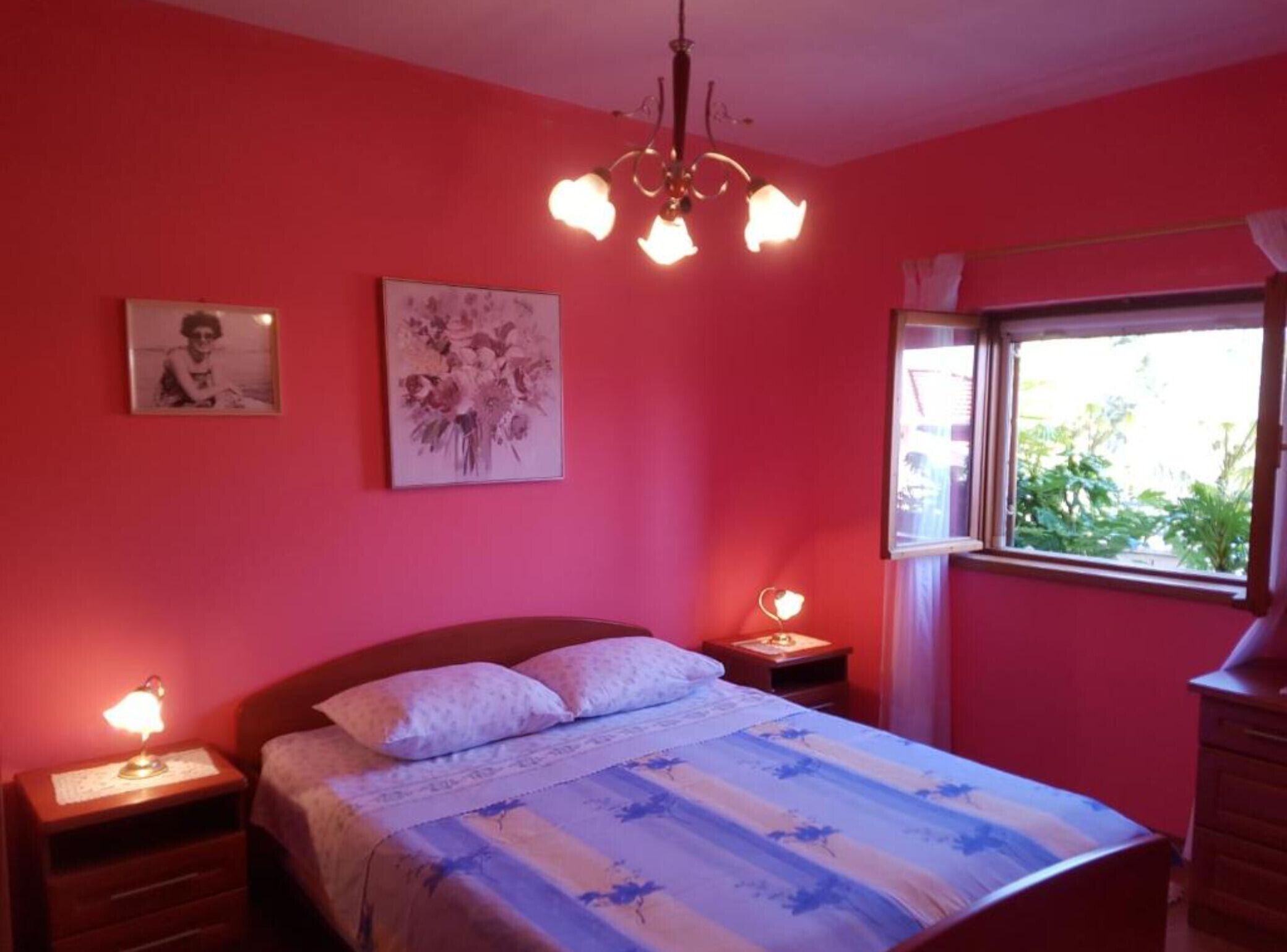 A bedroom with a bed, two bedside tables, a chandelier, a window with a view of greenery, and two wall-mounted pictures.