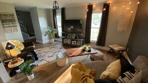 Una sala de estar acogedora con un sofá, una mesa de centro y un televisor. Se ven plantas, una lámpara y una escalera al fondo.