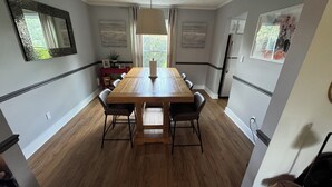 Un comedor con una mesa y sillas de madera, una mesita auxiliar roja con fotos enmarcadas y un ventanal con cortinas.