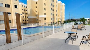 Una piscina cercada, con mobiliario exterior y un edificio de varios pisos al fondo.
