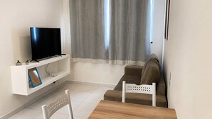 Una sala de estar con un televisor sobre un estante blanco, un sofá marrón, una mesa de madera y una silla blanca.
