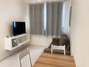 Uma sala de estar com uma TV em uma estante branca, um sofá marrom, uma mesa de madeira e uma cadeira branca.