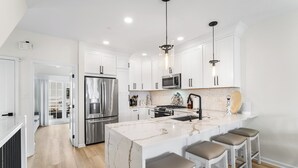 Una cocina moderna con armarios blancos, una isla grande con taburetes, electrodomésticos de acero inoxidable e iluminación colgante.