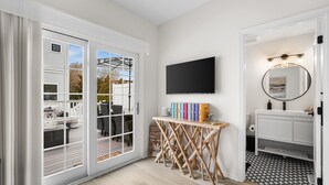 Una habitación con un gran ventanal corredero que da a un espacio exterior, un televisor de pantalla plana montado en la pared, una mesita de madera con libros y un tocador blanco con espejo e iluminación.