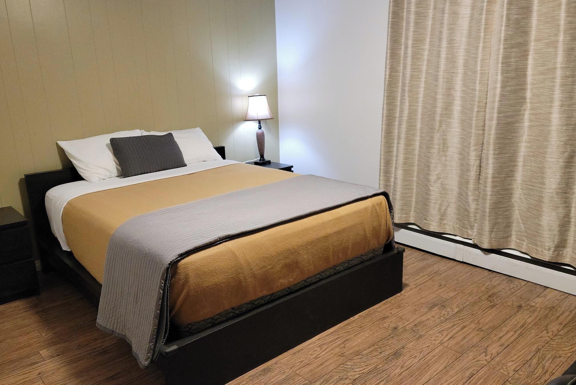 A neatly made bed with a beige and gray blanket, a black nightstand with a lamp, and a wooden floor.