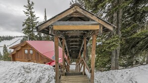 Una escalera de madera lleva a un chalé con techo rojo, rodeado de nieve y árboles.