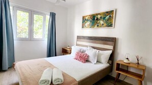 A neatly made bed with white and pink pillows, a wooden headboard, and a pair of towels on the bed. There is a small bedside table with a lamp and a decorative item. A painting hangs on the wall above the bed. - Apartamento Bayahibe Con Terraza (San Rafael del Yuma)