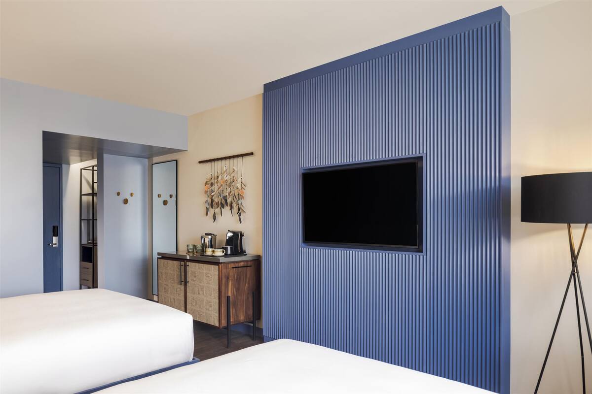 Une chambre d’hôtel avec un grand téléviseur fixé sur un mur bleu, une commode en bois, un lit et un miroir.