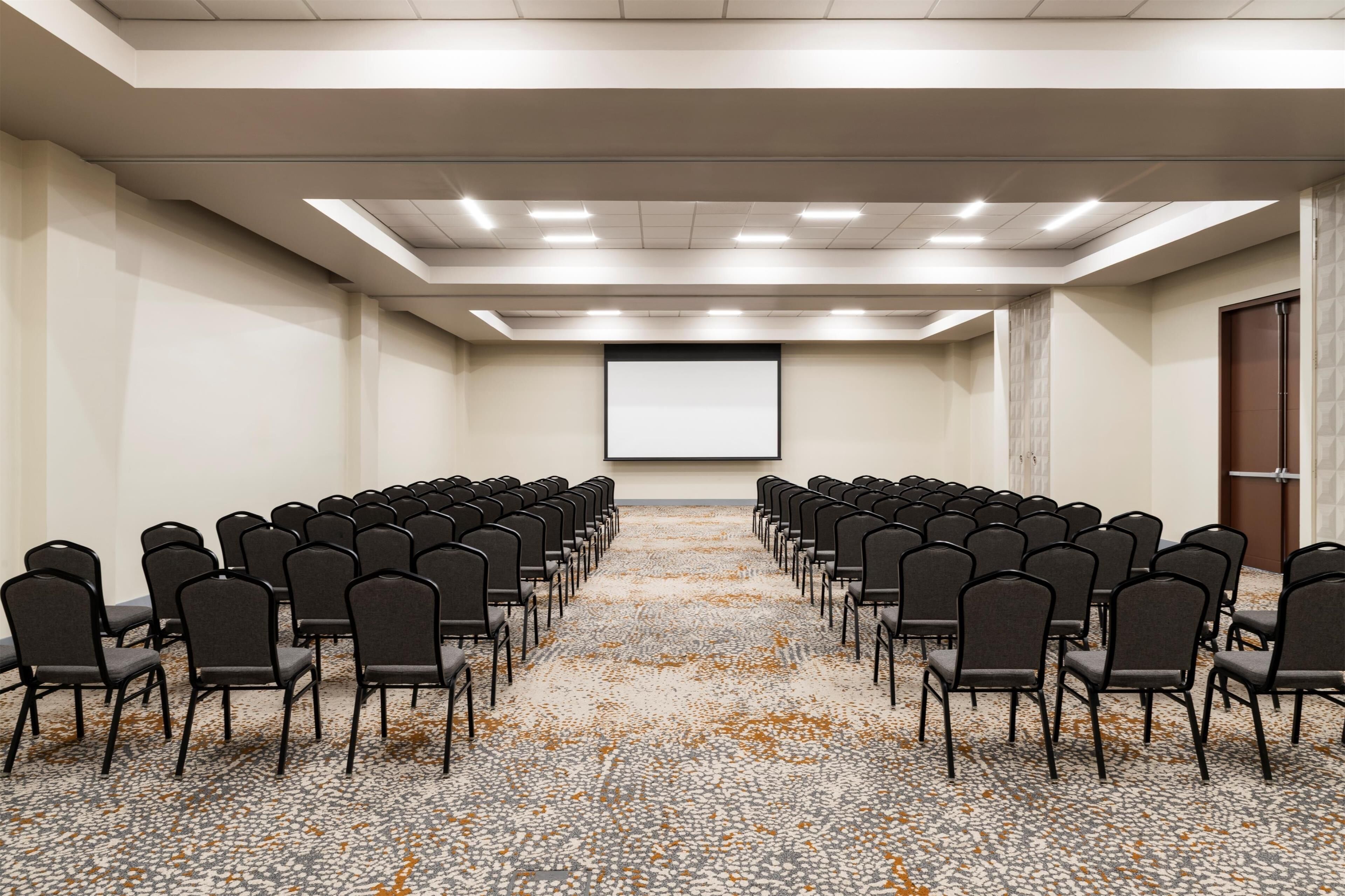 A conference room with rows of chairs facing a projection screen, carpeted floor, and a door on the right.