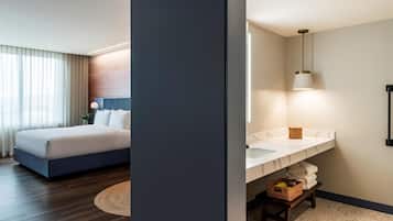 Habitación de hotel con cama, mesita de noche, lámpara y baño con lavamanos, espejo, toallero y un taburete pequeño.