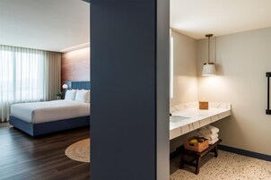 Habitación de hotel con cama, mesita de noche, lámpara y baño con lavamanos, espejo, toallero y un taburete pequeño.