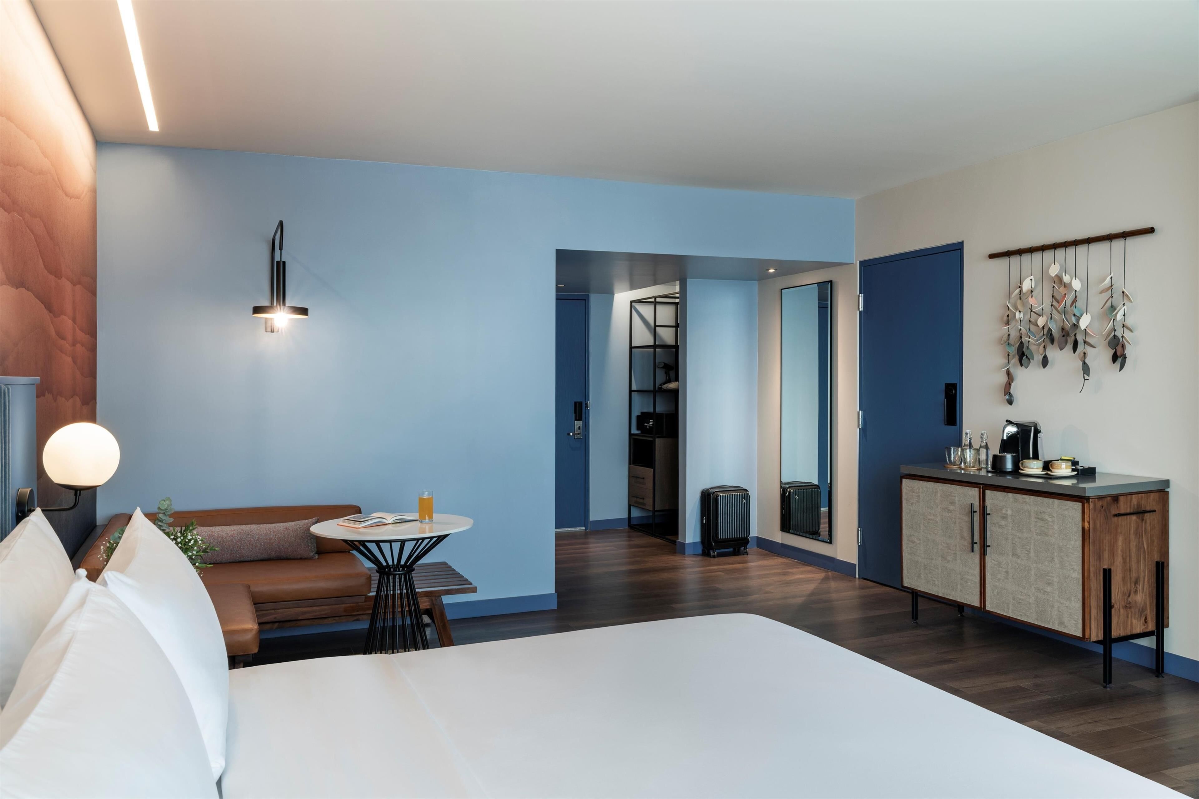 Een moderne hotelkamer met een bed, een klein rond tafeltje, een bank, een kitchenette en een badkamer zichtbaar door een geopende deur.