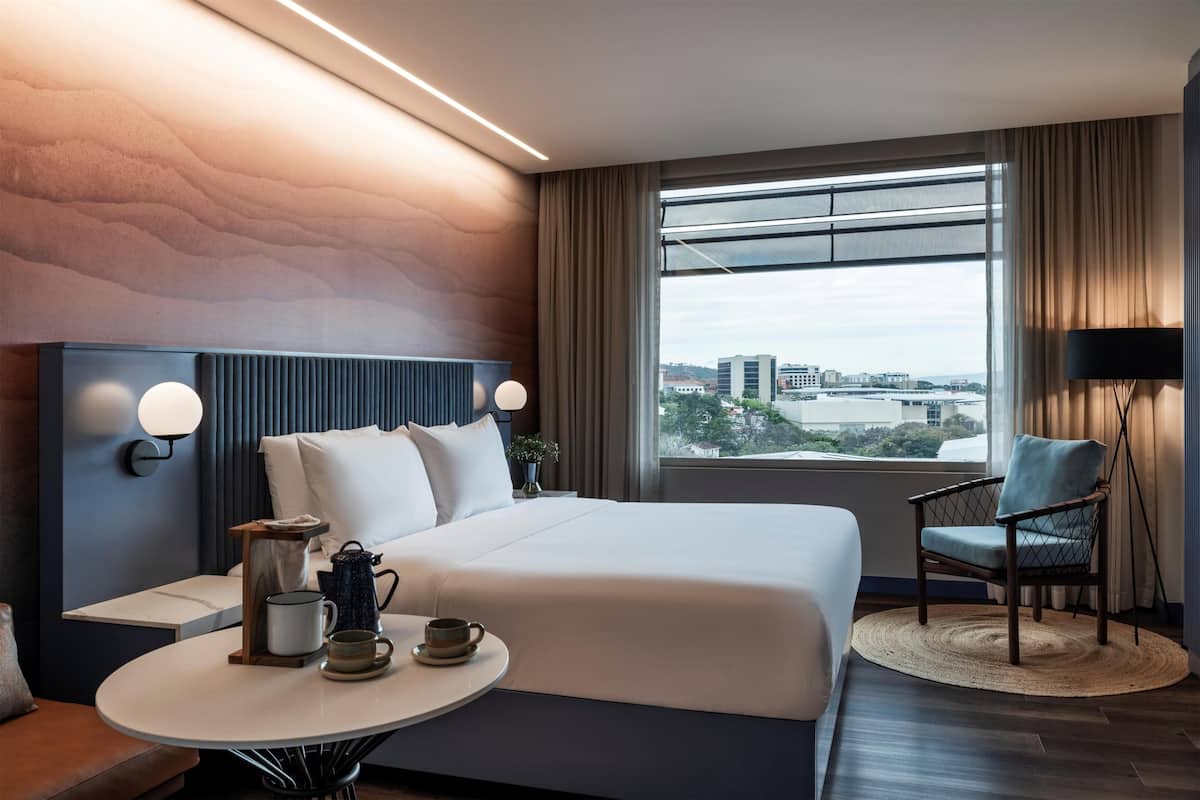 Une chambre d’hôtel moderne dotée d’un grand lit, d’une table ronde avec un plateau pour le café, d’une chaise et offrant une vue sur la ville grâce à de grandes fenêtres.