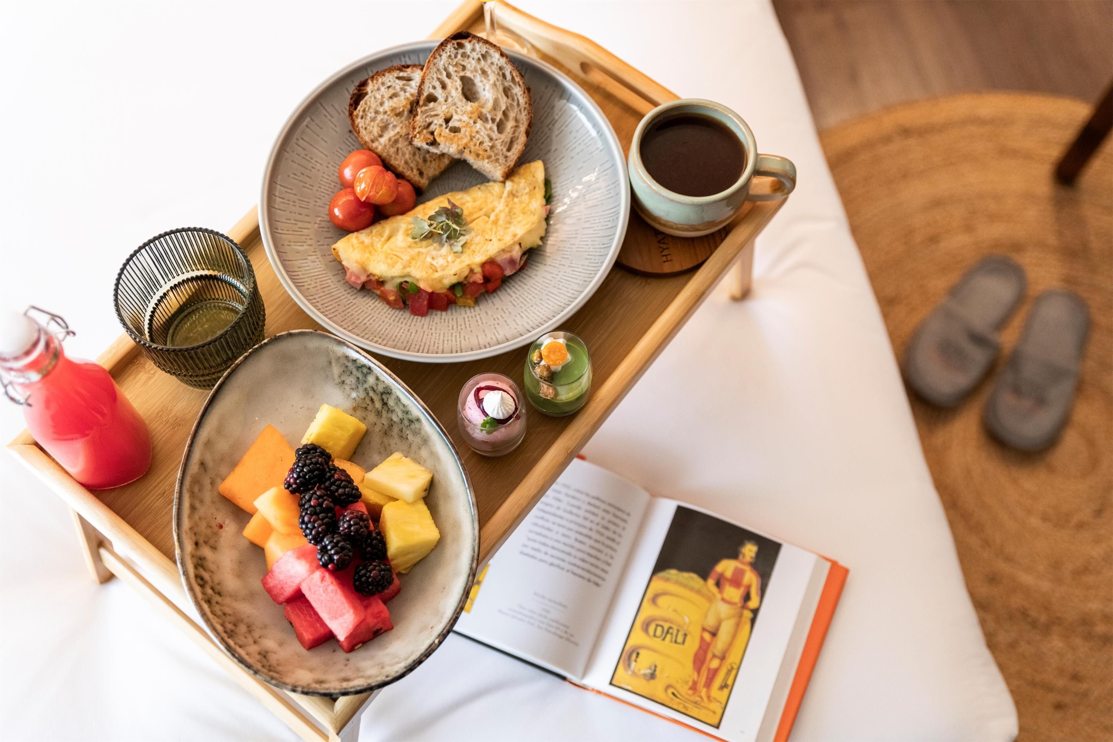 Una bandeja de desayuno con tostadas, huevos, tomates y una taza de café sobre una cama con pantuflas y un libro.