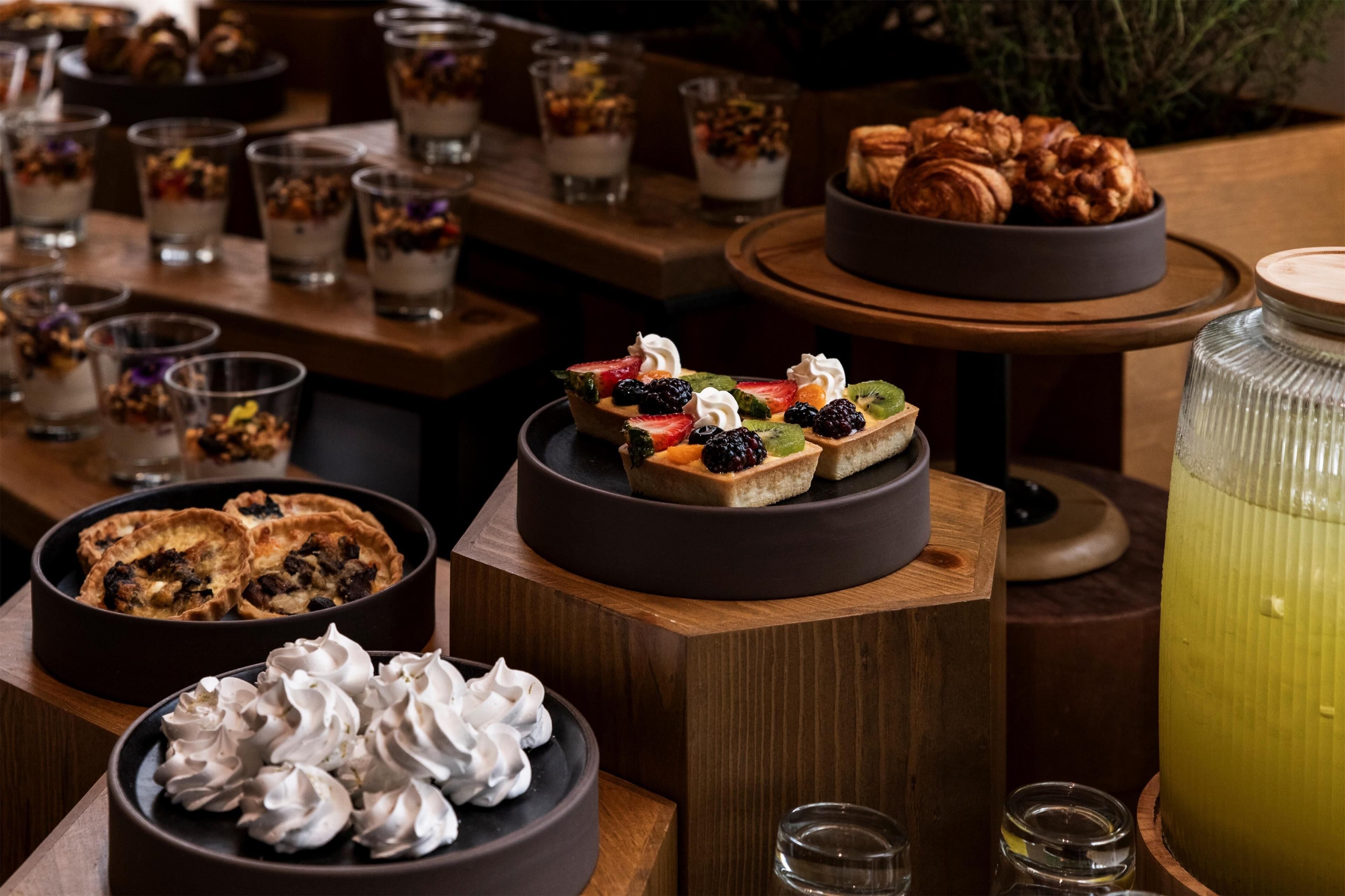 A variety of desserts and beverages on wooden tables, including cakes, tarts, and a large jar of lemonade.