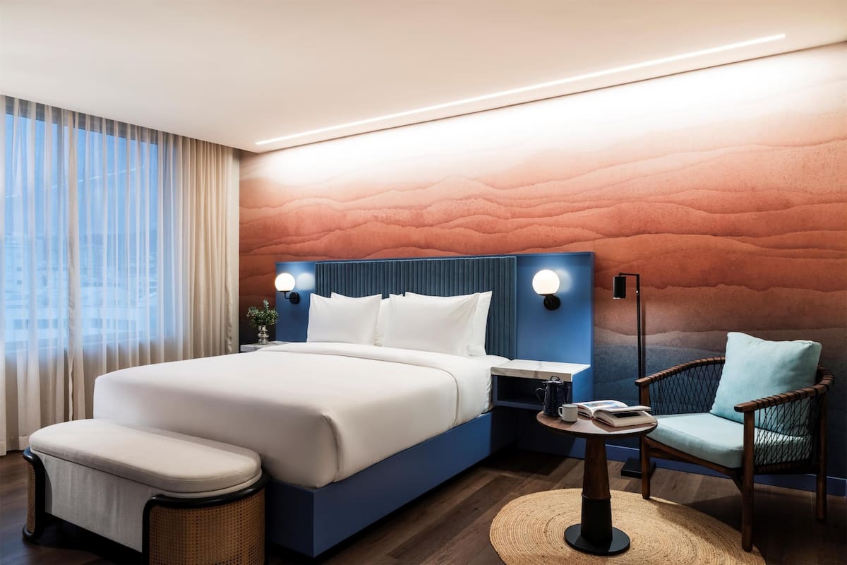 Une chambre d’hôtel moderne dotée d’un grand lit, d’un coin salon avec un fauteuil et une petite table, ainsi que d’une table de chevet avec une lampe.