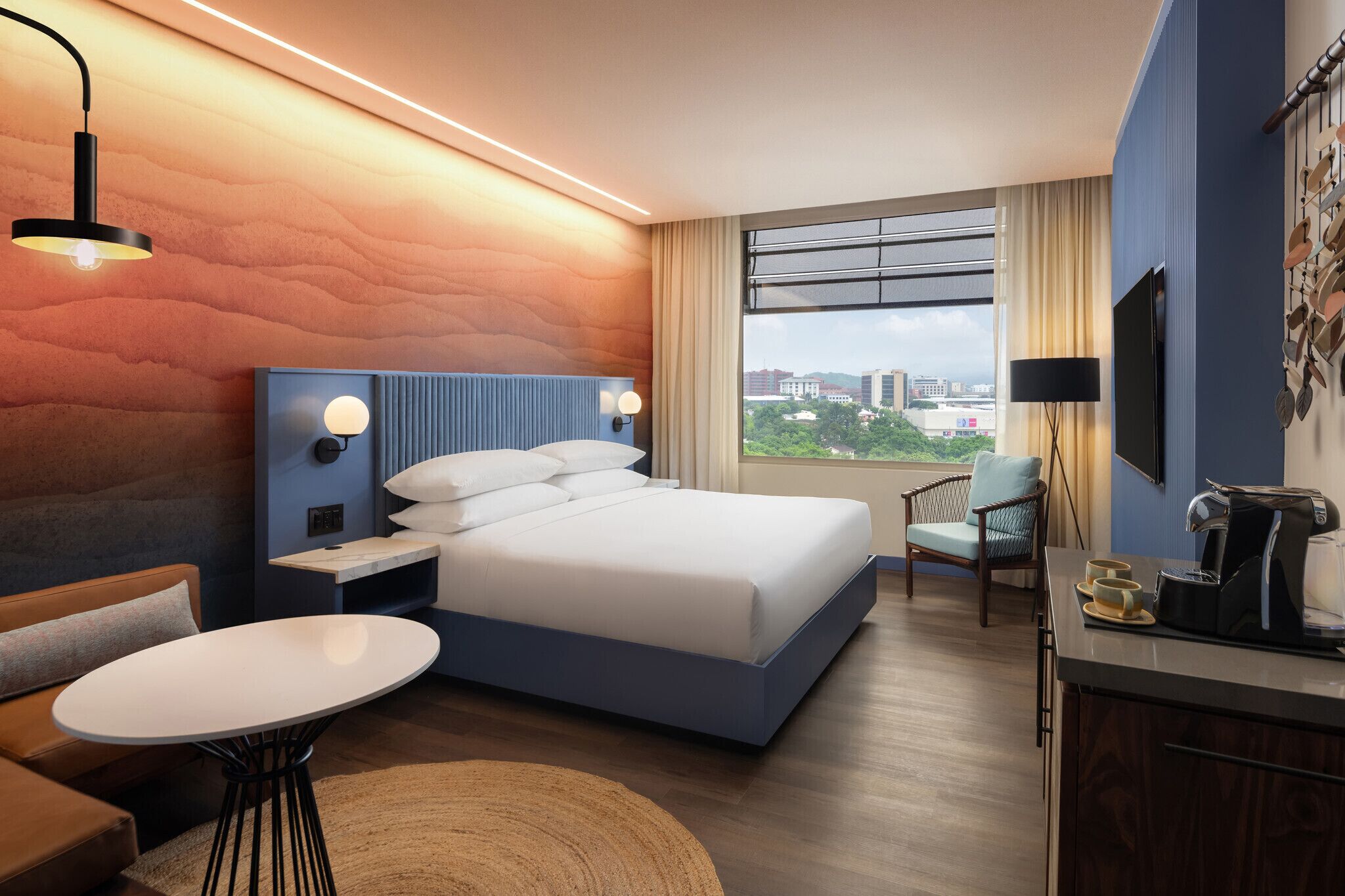A modern hotel room with a large bed, a small table, a chair, a desk with a coffee maker, and a TV mounted on the wall.