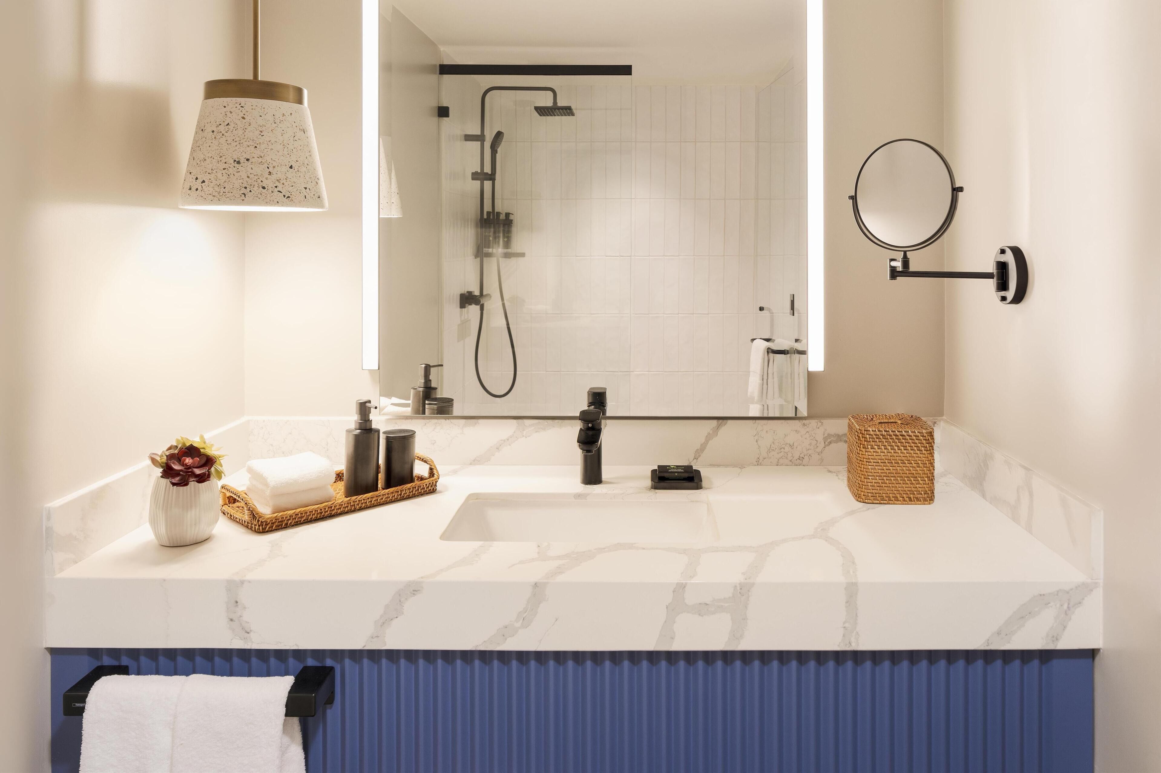 A modern bathroom with a marble sink, a handheld showerhead, a round mirror, and a towel rack.