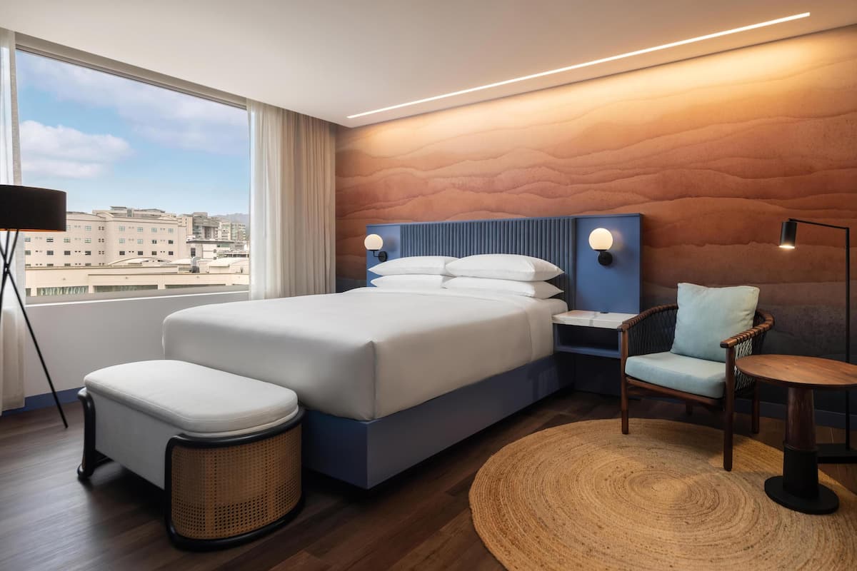 Une chambre d’hôtel moderne, dotée d’un grand lit, d’une table de chevet, d’une chaise, d’une table ronde et offrant une vue sur la ville grâce à de grandes fenêtres.