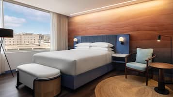 Ein modernes Hotelzimmer mit einem großen Bett, einem Nachttisch, einem Sessel, einem runden Tisch und Blick auf die Stadt durch große Fenster.