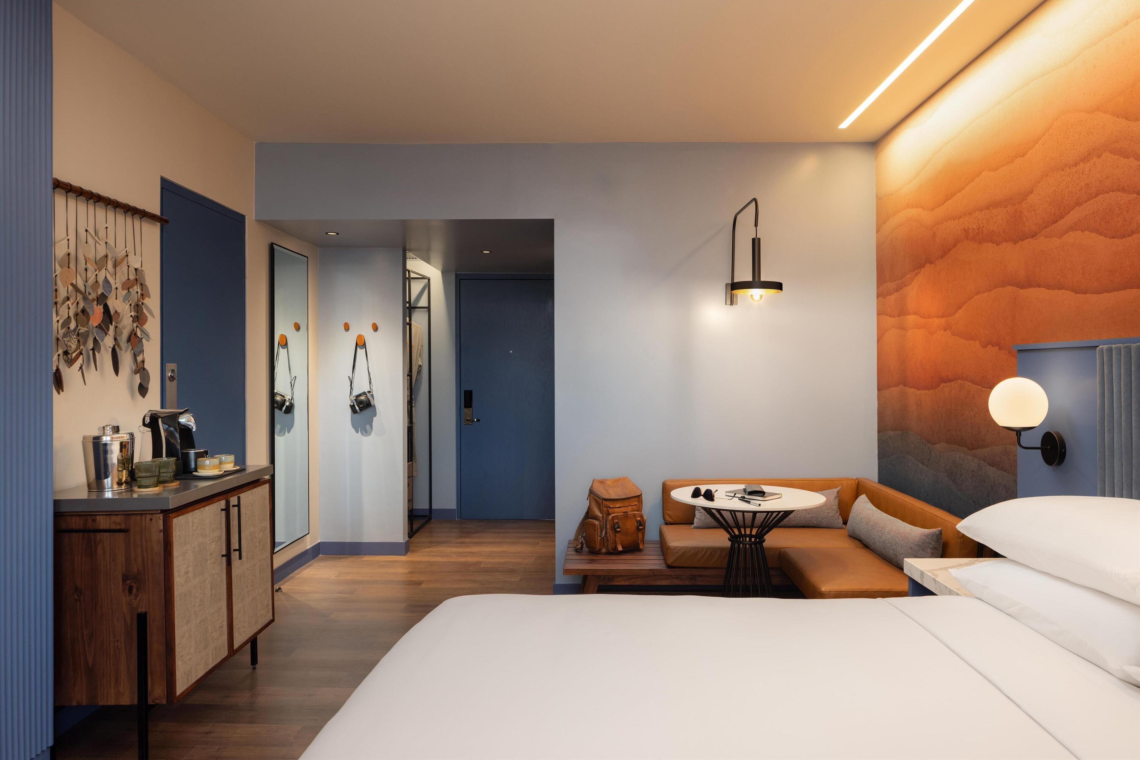 Ein modernes Hotelzimmer mit Holzboden, einem Bett mit weißer Bettwäsche, einer Sitzecke mit Tisch und einer Küchenzeile.