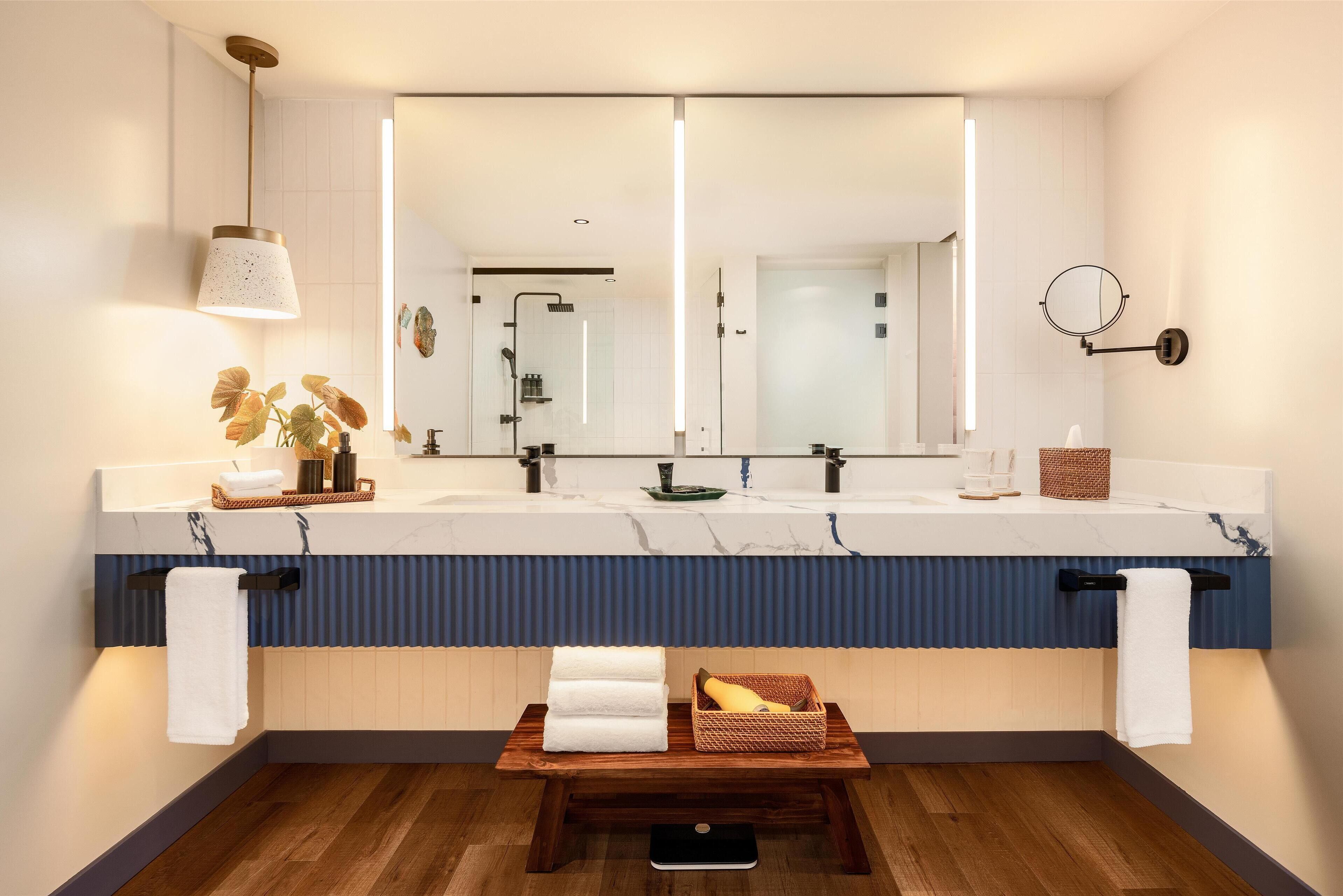 A modern bathroom with a large mirror, two sinks, a wooden bench with towels, and a plant.