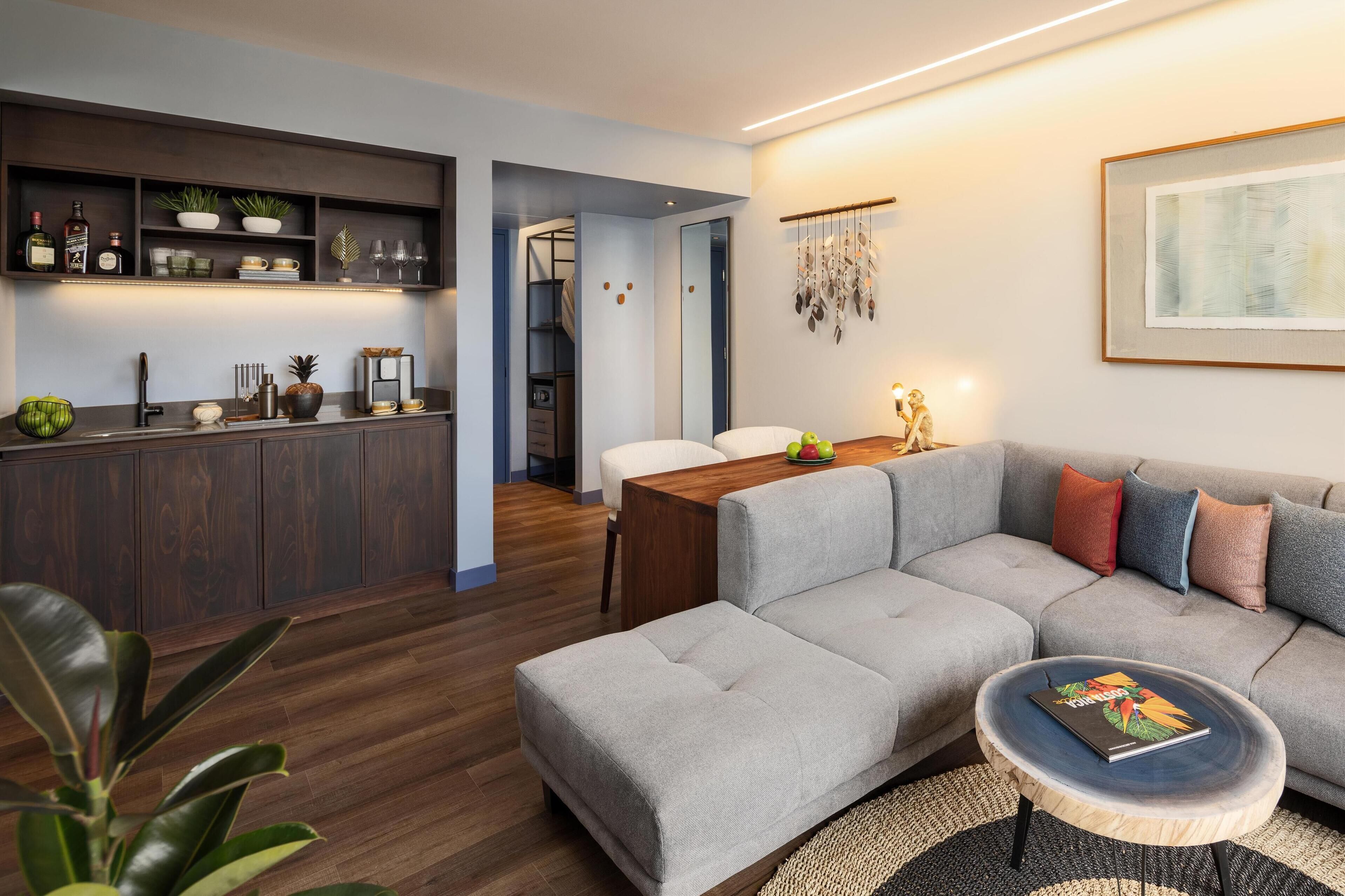 Un salon moderne avec un canapé d’angle gris, une table basse en bois et un coin cuisine doté d’un meuble et d’étagères en bois foncé.