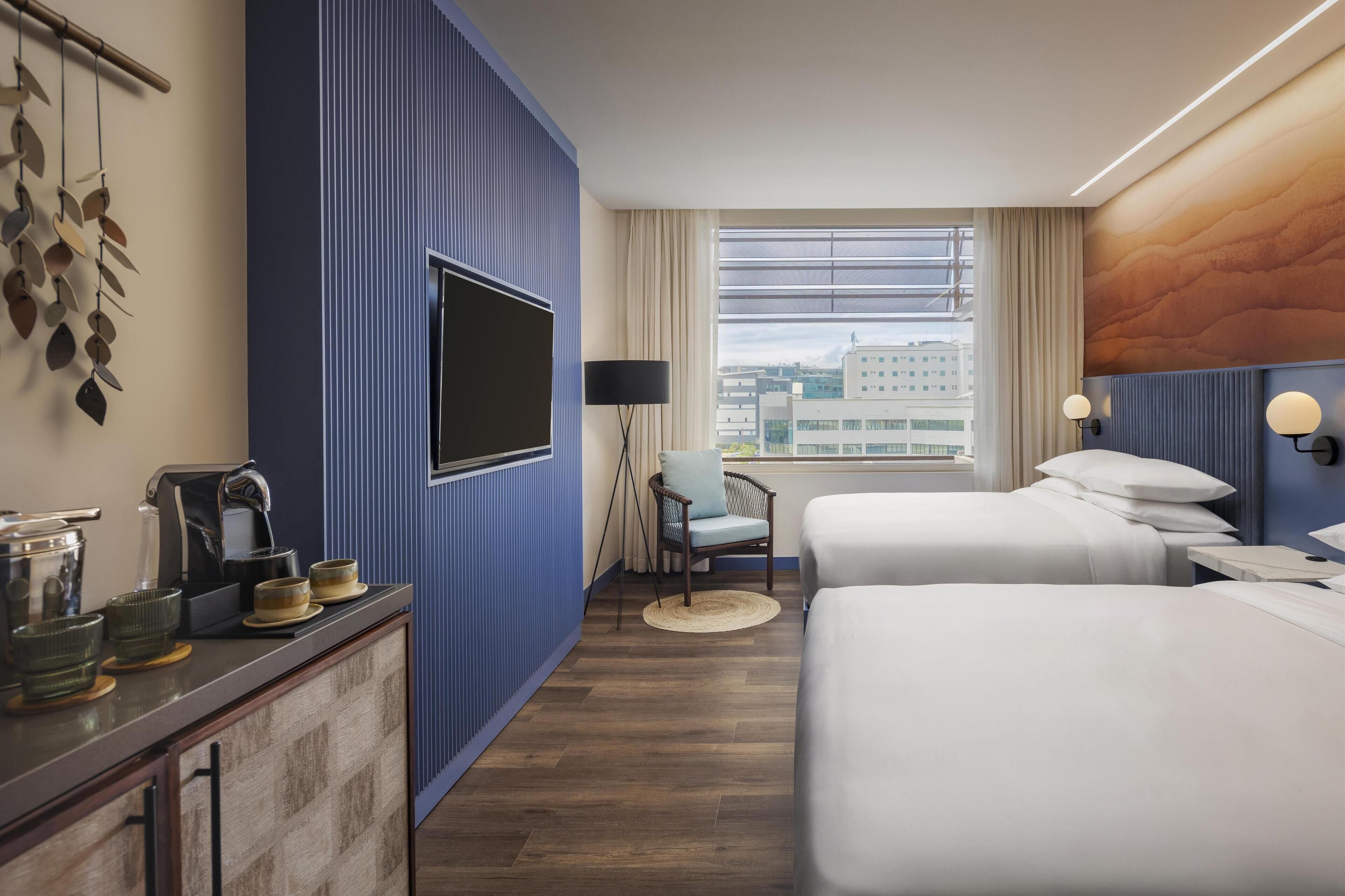 A hotel room with two beds, a TV, a desk with a coffee maker, and a view of the city through large windows.