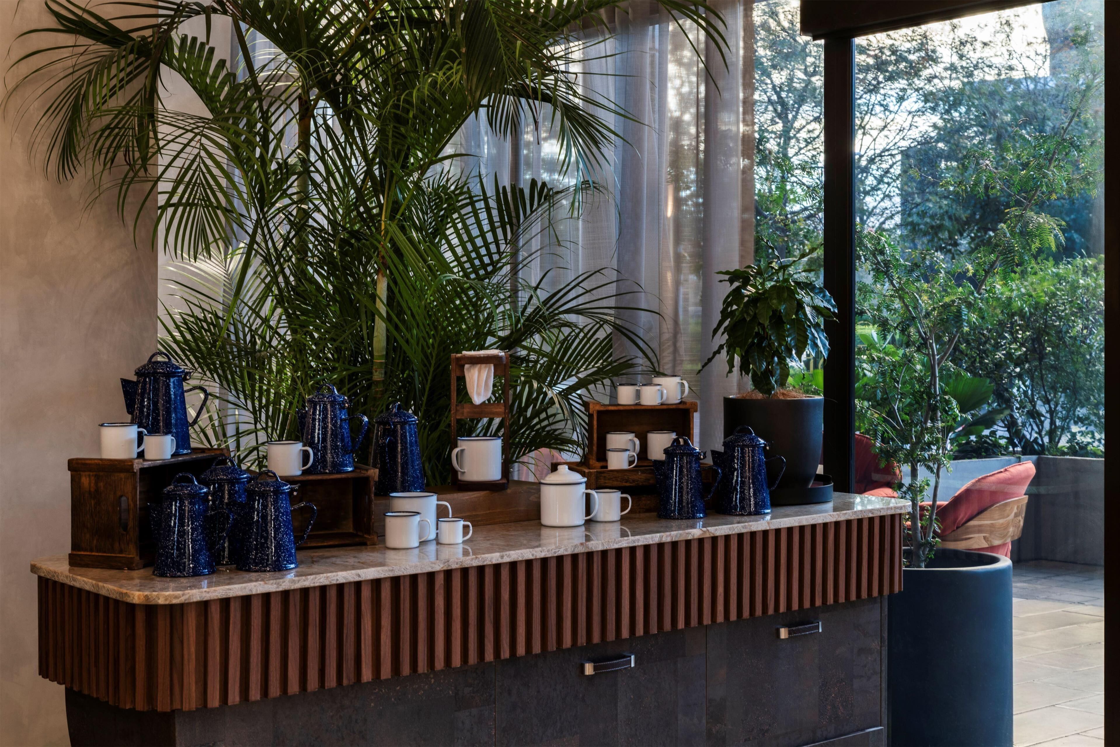 A counter with various coffee pots and mugs, surrounded by large potted plants.