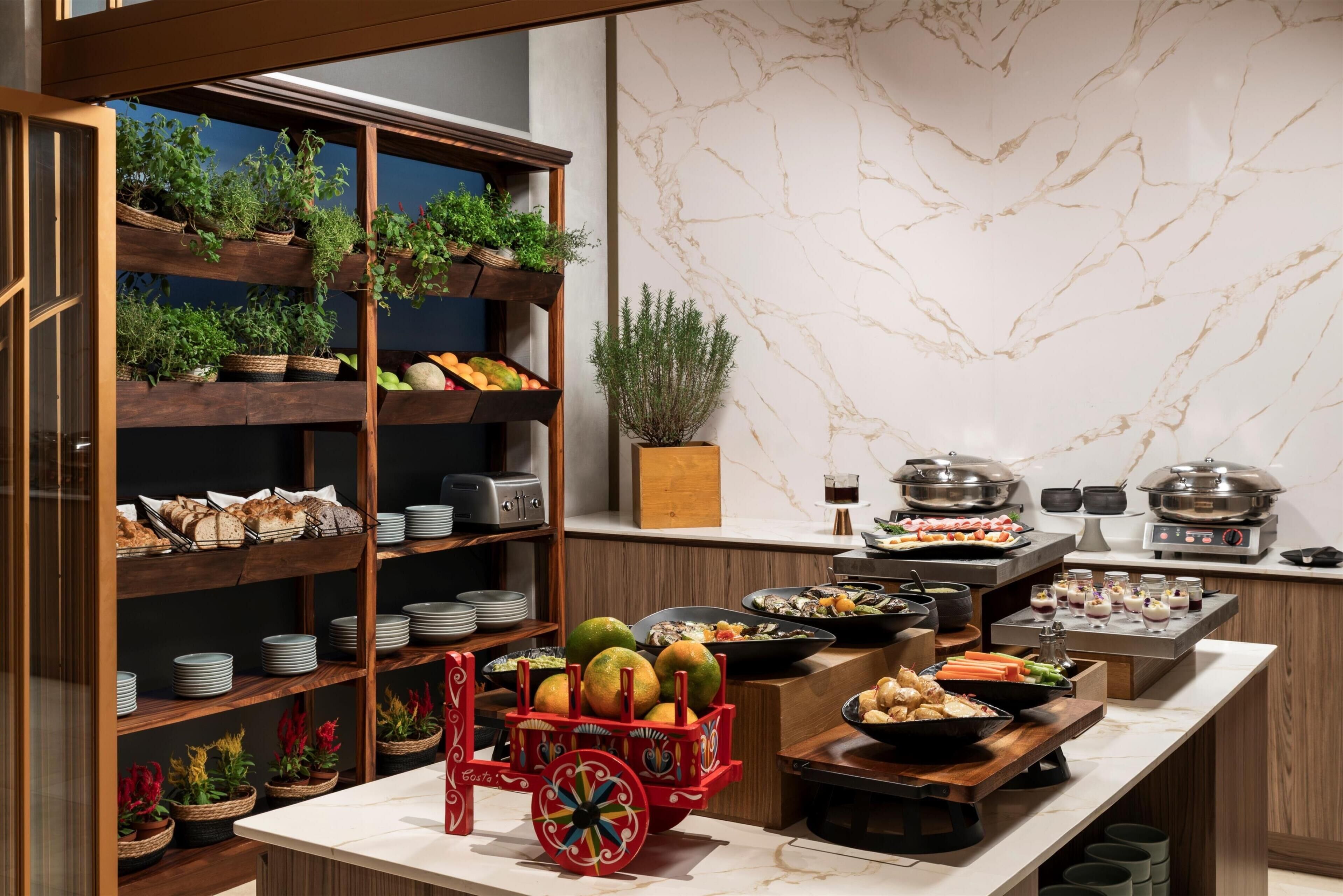 A buffet setup with various food items, a fruit cart, and shelves with baskets of plants and bread.