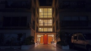 A multi-story building with a central entrance, a car parked to the right, and a well-lit facade.