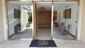 Entrada de un hotel con puerta de cristal, mostrador de recepción y zona de espera con sillas.