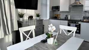 A modern kitchen with a flat-screen TV, microwave, and stainless steel range hood. There are two white chairs and a dining table with a vase of flowers. - Granada Apartments Holmfield Rd flat 3 1st floor front (Blackpool)