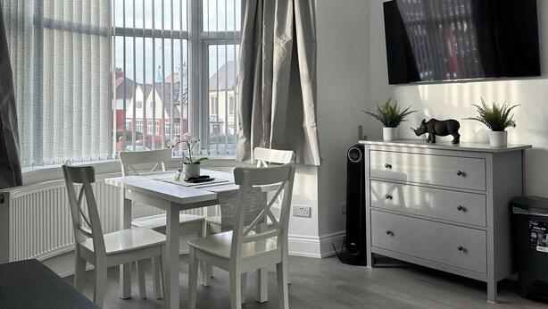 A modern, well-lit room with a dining table, chairs, a TV mounted on the wall, and a radiator. - Granada Apartments Holmfield Rd flat 3 1st floor front (Blackpool)
