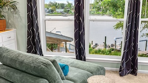 Un sofá verde con un cojín azul, una pequeña mesa redonda de madera y vistas a una masa de agua con un muelle y vegetación.