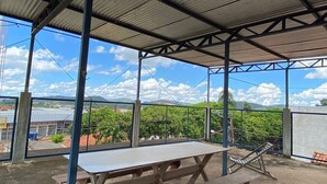 A covered outdoor area with a picnic table and benches, surrounded by a metal fence and a view of green vegetation and distant buildings. - Hotel ferraz (Tucumã)