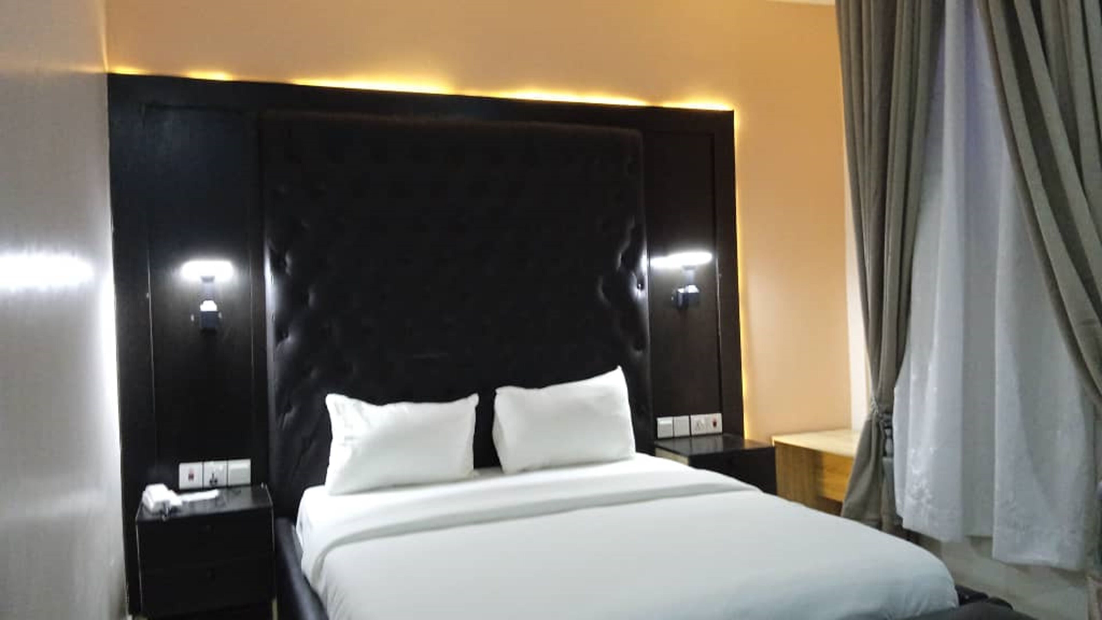 Habitación de hotel con una cama grande, cabecera oscura, lámparas de noche y cortina en la ventana.