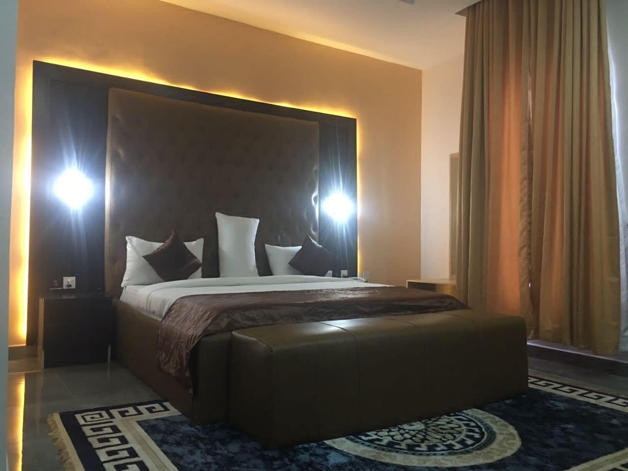 Una habitación de hotel moderna con una cama grande, cabecera con iluminación integrada, mesita de noche y una alfombra estampada.